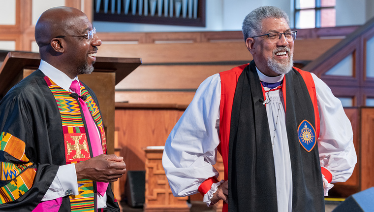 Two clergy members in robes stand inside a church, with wooden paneling and an organ in the background.