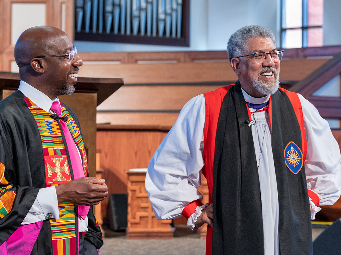 Two clergy members in robes stand inside a church, with wooden paneling and an organ in the background.