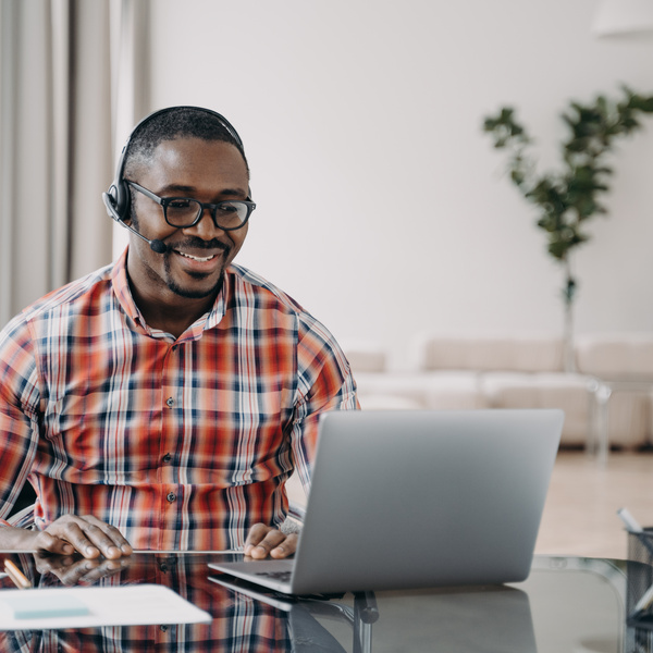 Smiling african american young man wearing wireless headset with microphone, looking at laptop screen, learning online. Professional male teacher giving lesson to client, working remotely. E-learning.