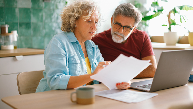 Senior husband and wife paying bills and managing family finances together on a laptop, discussing expenses in a cozy home environment.