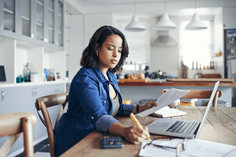 Shot of a young woman using a laptop and  going through paperwork while working from home